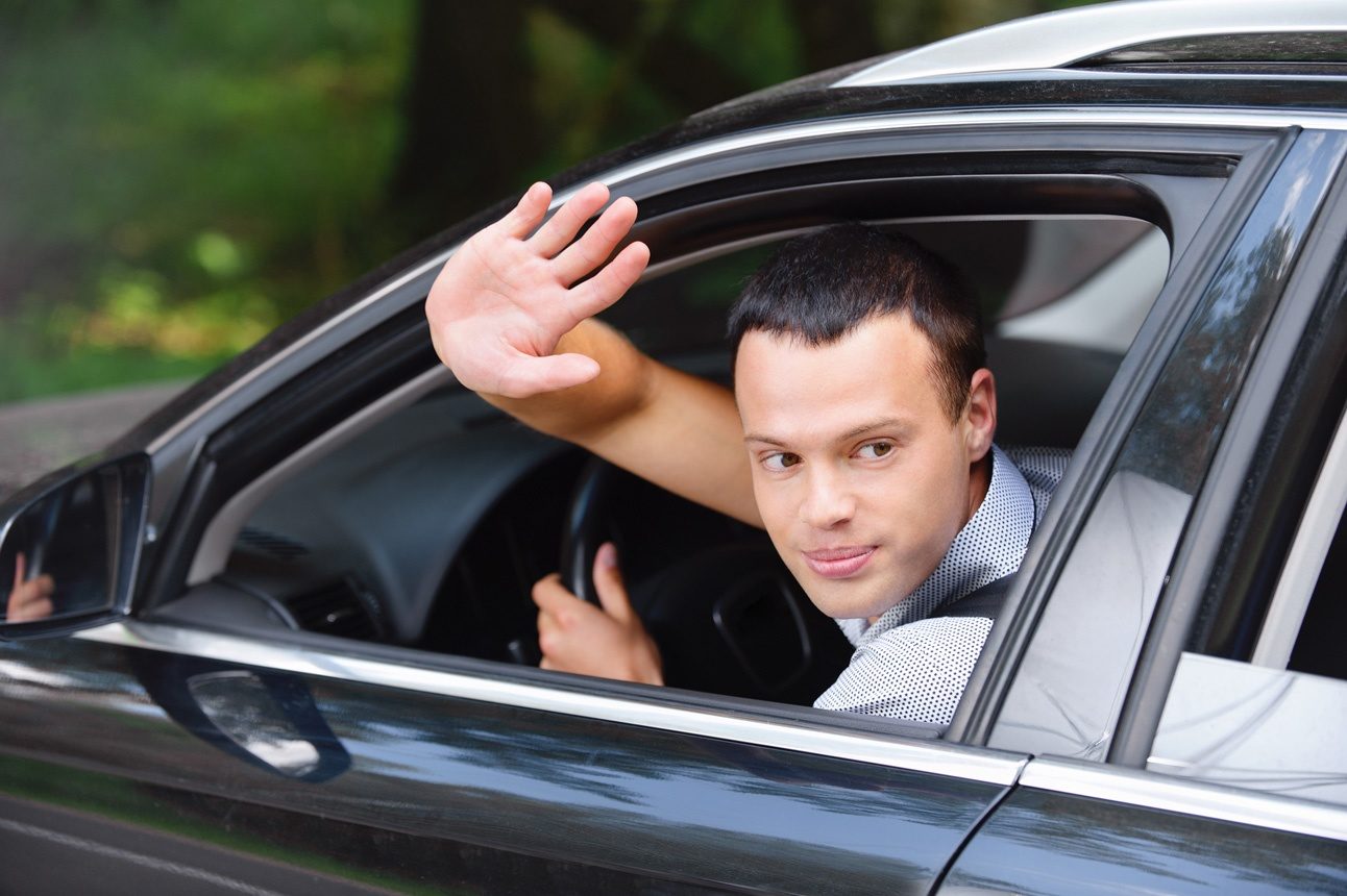 Un jeune homme au volant de sa voiture en train de saluer de la main pour dira à la fois merci et désolé.