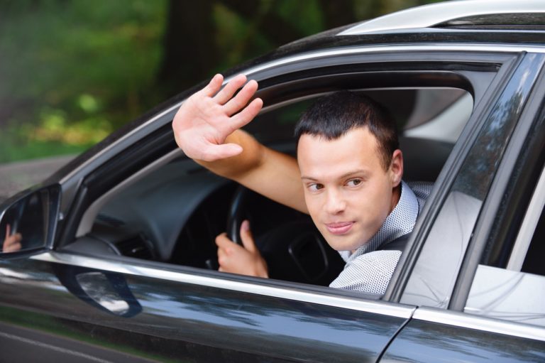 Un jeune homme au volant de sa voiture en train de saluer de la main pour dira à la fois merci et désolé.