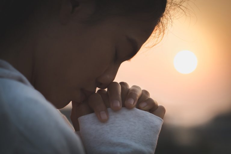 Une jeune femme en train de prier, les deux mains jointes, en extérieur et à l'aube.
