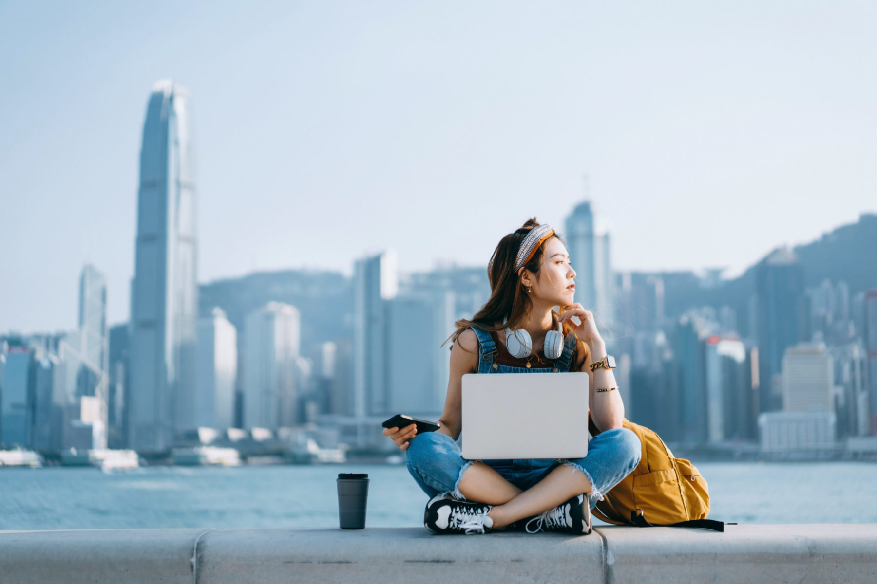 Une jeune femme assise en tailleur avec un laptop regarde sur le côté d'un air songeur. A l'arrière, une grande ville.