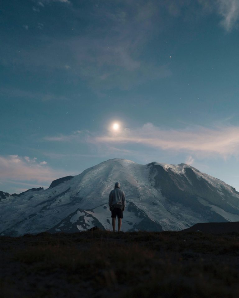 Une personne se tient debout à l'extérieur, dans la nuit éclairée par la lune, devant une montagne.