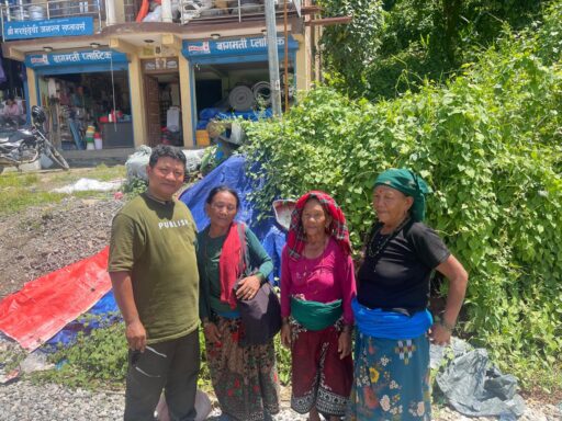 Un homme (Prem Tamang) vêtu d'un t-shirt vert pose aux côtés de trois femmes népalaises âgées, habillées de tenues traditionnelles colorées et de foulards. Le groupe se tient à l'extérieur sur un sol rocailleux, devant des bâches en plastique de fortune, une végétation touffue et la devanture de petits commerces locaux.