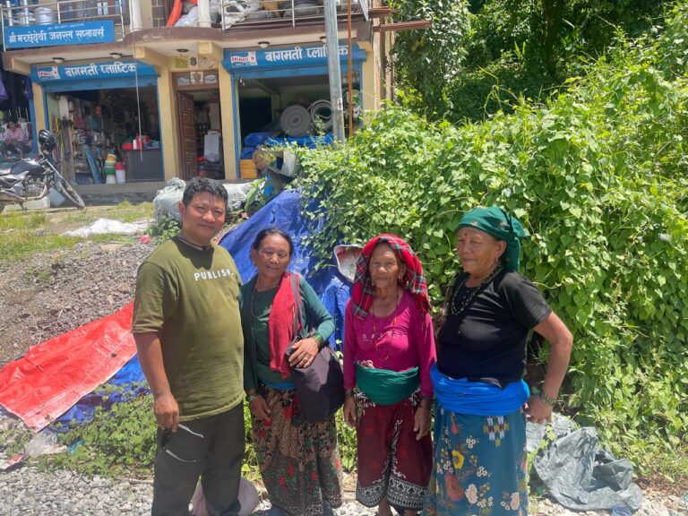 Un homme (Prem Tamang) vêtu d'un t-shirt vert pose aux côtés de trois femmes népalaises âgées, habillées de tenues traditionnelles colorées et de foulards. Le groupe se tient à l'extérieur sur un sol rocailleux, devant des bâches en plastique de fortune, une végétation touffue et la devanture de petits commerces locaux.