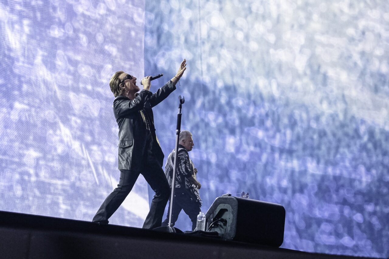 Bono sur scène, micro en main et bras levé, lors d'un concert de U2